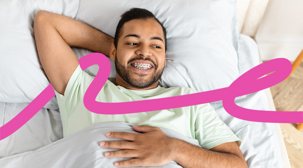 Smiling man with braces lying in bed under a blanket with a bold pink ribbon graphic across the scene.