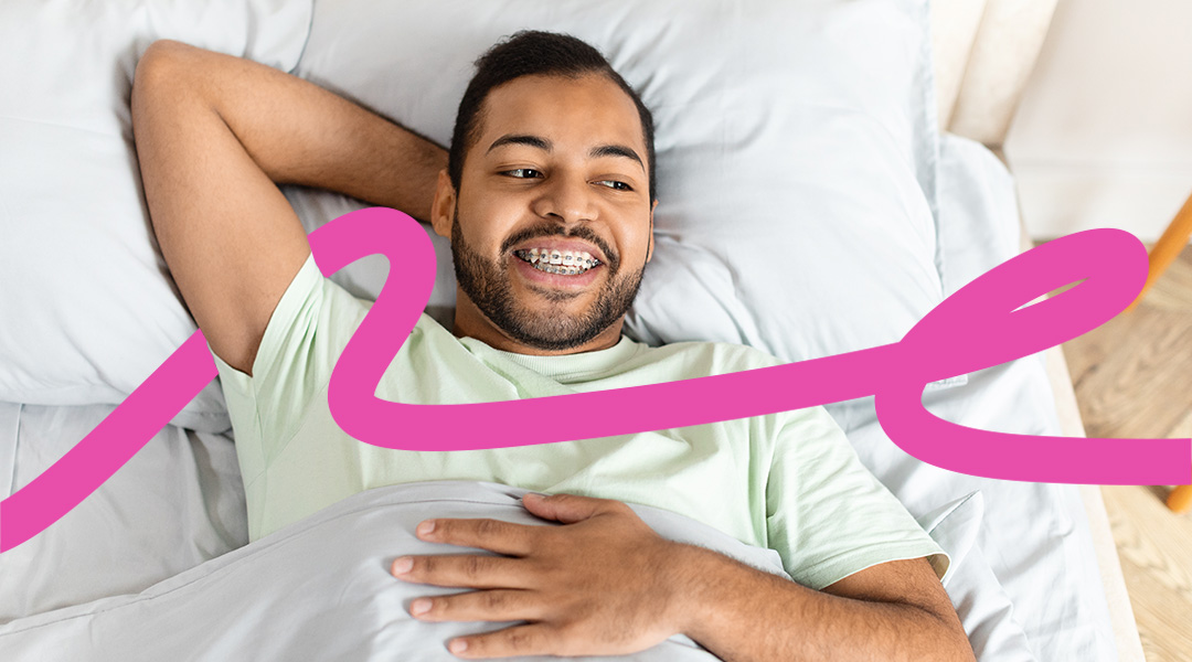 Smiling man with braces lying in bed under a blanket with a bold pink ribbon graphic across the scene.