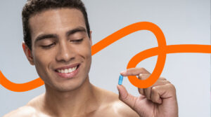 Smiling man holds a blue capsule between his fingers against a light gray background with an orange decorative loop behind him.